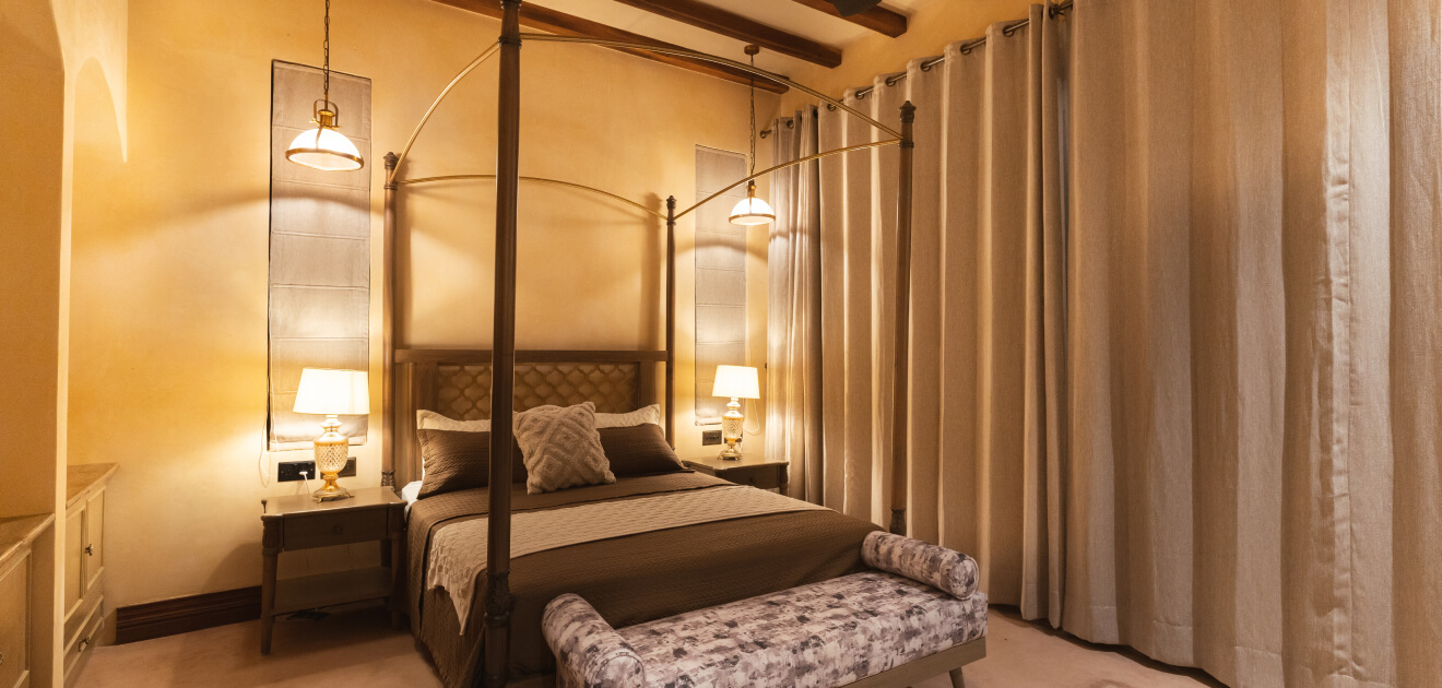 A cozy bedroom featuring a four-poster bed with beige and patterned linens, ambient lighting, wooden ceiling beams, and draped curtains around the room.