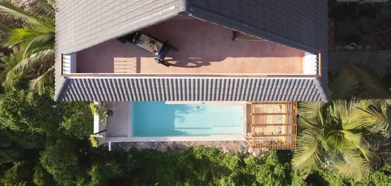 Aerial view of a symmetrical backyard with pool, roof , surrounded by green foliage, at dusk in a house.