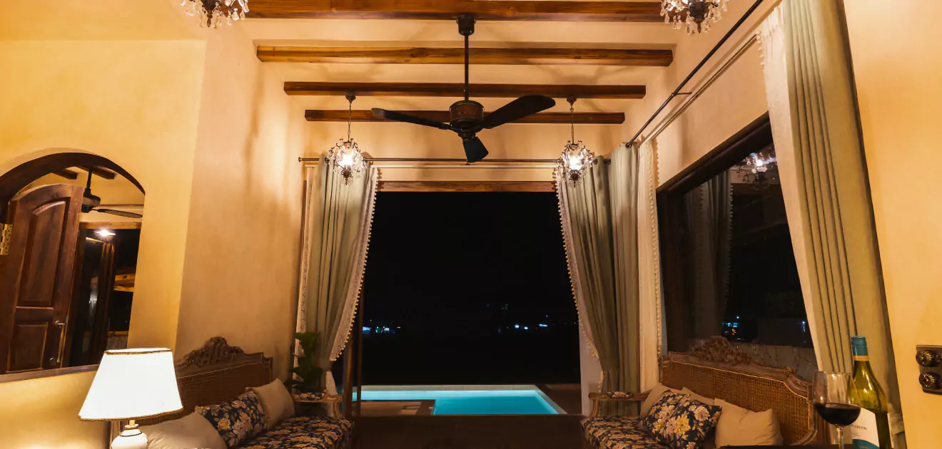 An elegant room with a pool view through an open door, featuring warm lighting, plush seating, wooden ceiling beams, and a ceiling fan.