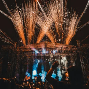 A lively concert with a crowd and fireworks bursting above the stage at night.