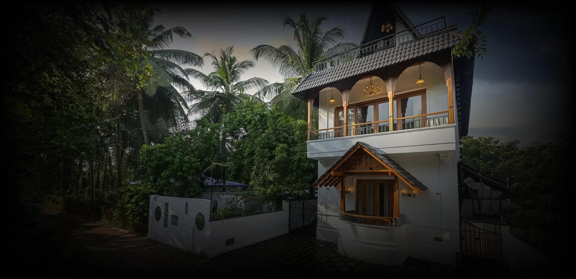 A modern three-story house illuminated at twilight, featuring large windows, balconies, and a pool, set against a backdrop of trees and a sunset sky.